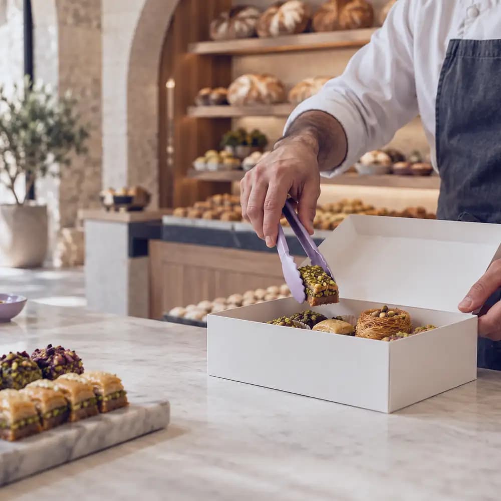 A baker boxes sweets with tongs at a bakery counter.