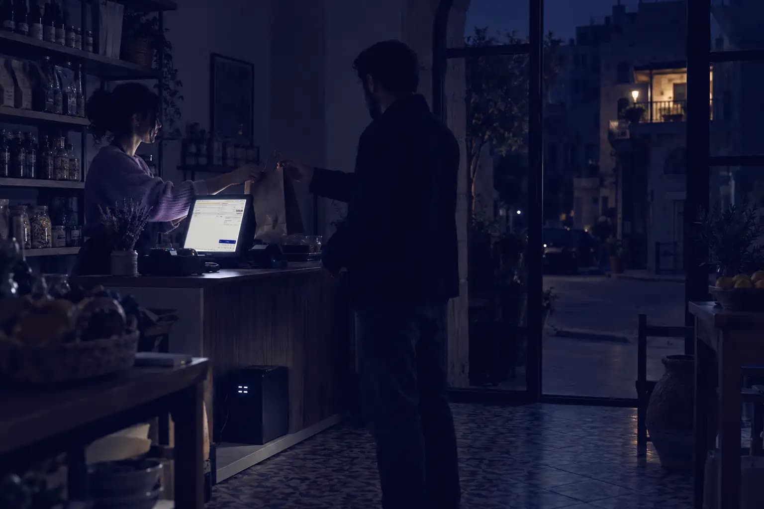 A shop at dusk during a power cut: the room is dark, and only the counter screen glows while the cashier serves a customer.